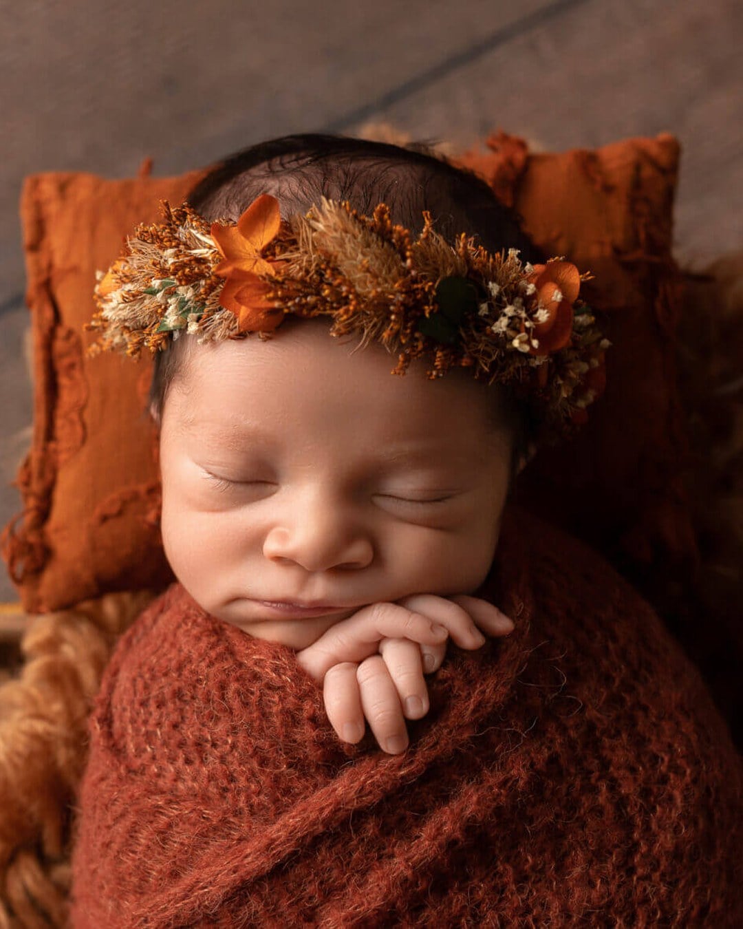 A sleeping newborn in a rust knit wrap wearing a dried-flower crown, lying on a rust pillow and fleece in a bowl on a wooden floor