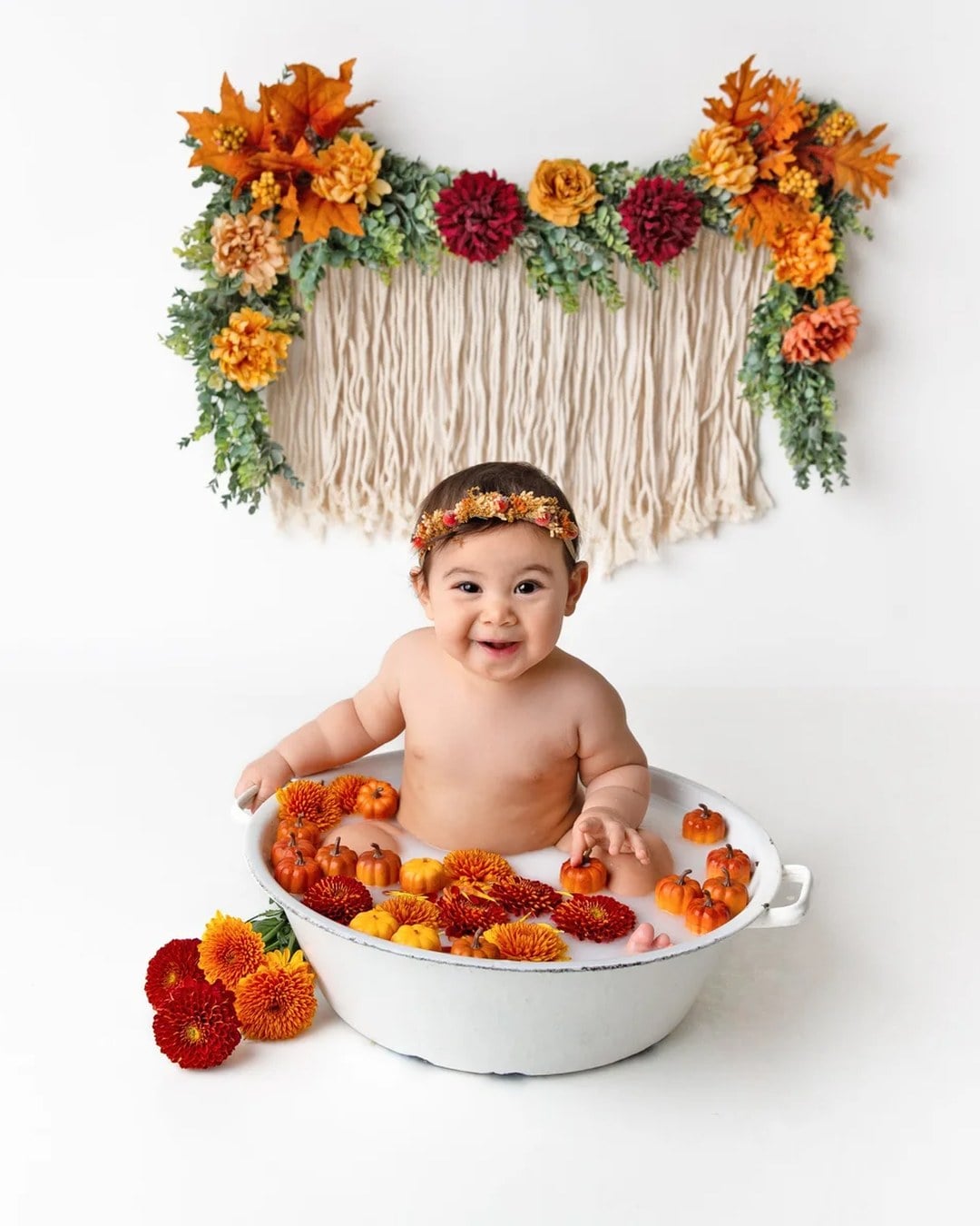 A smiling baby in a flower headband sitting in a white tub of milky water filled with small faux pumpkins and orange and red mums