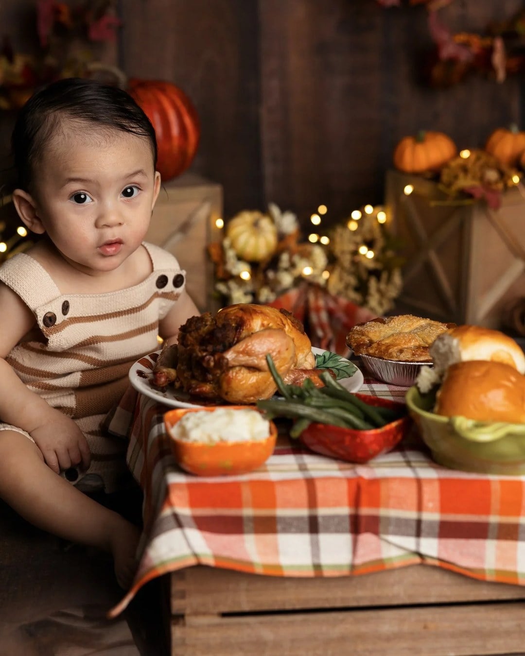 A baby sitting on a studio floor beside a crate table set with a roast turkey, mashed potatoes, green beans, rolls and a pie, with pumpkins and fairy lights behind