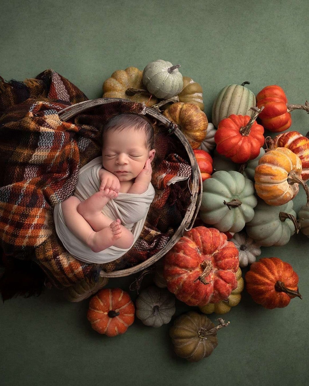 A sleeping newborn in a neutral wrap lying in a round basket on a plaid blanket among orange, teal and cream pumpkins on a green backdrop
