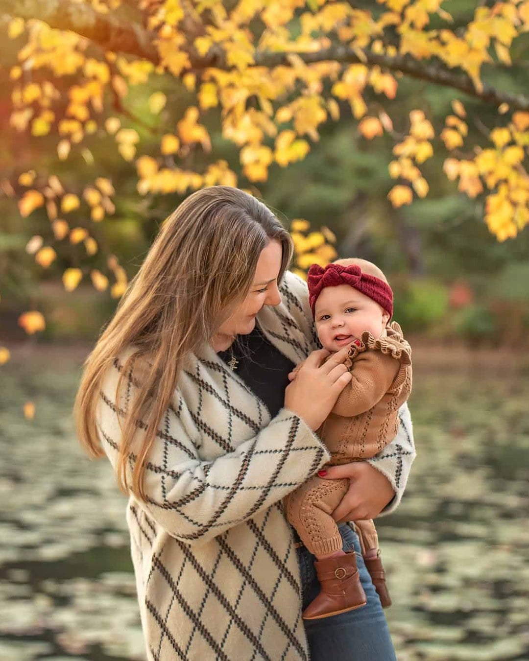 A baby in a berry-red knit headband and tan knit romper held in front of golden autumn leaves and a pond