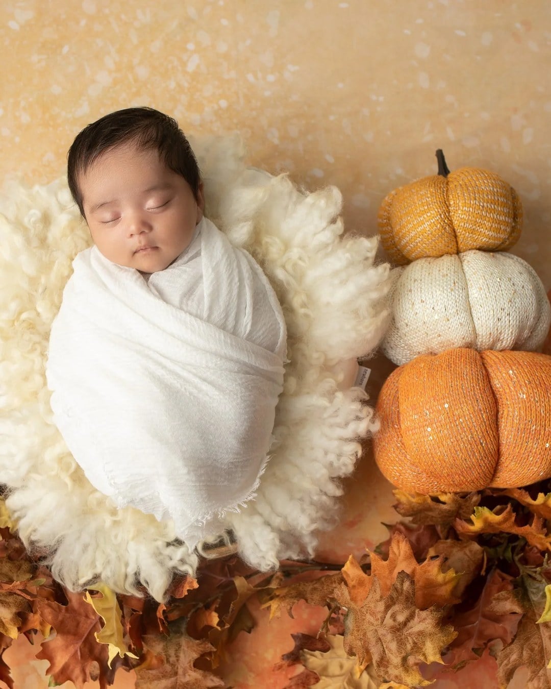 Overhead view of a sleeping newborn swaddled in white gauze on a cream fluffy rug with oak leaves, three stacked knit pumpkins and an orange backdrop