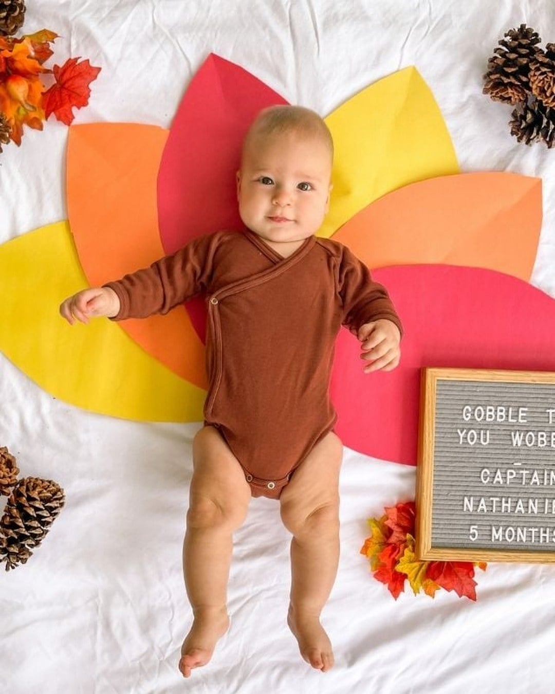 A baby in a brown onesie lying on a white sheet over paper turkey-feather petals with pinecones and a letterboard reading 'Gobble til you wobble'