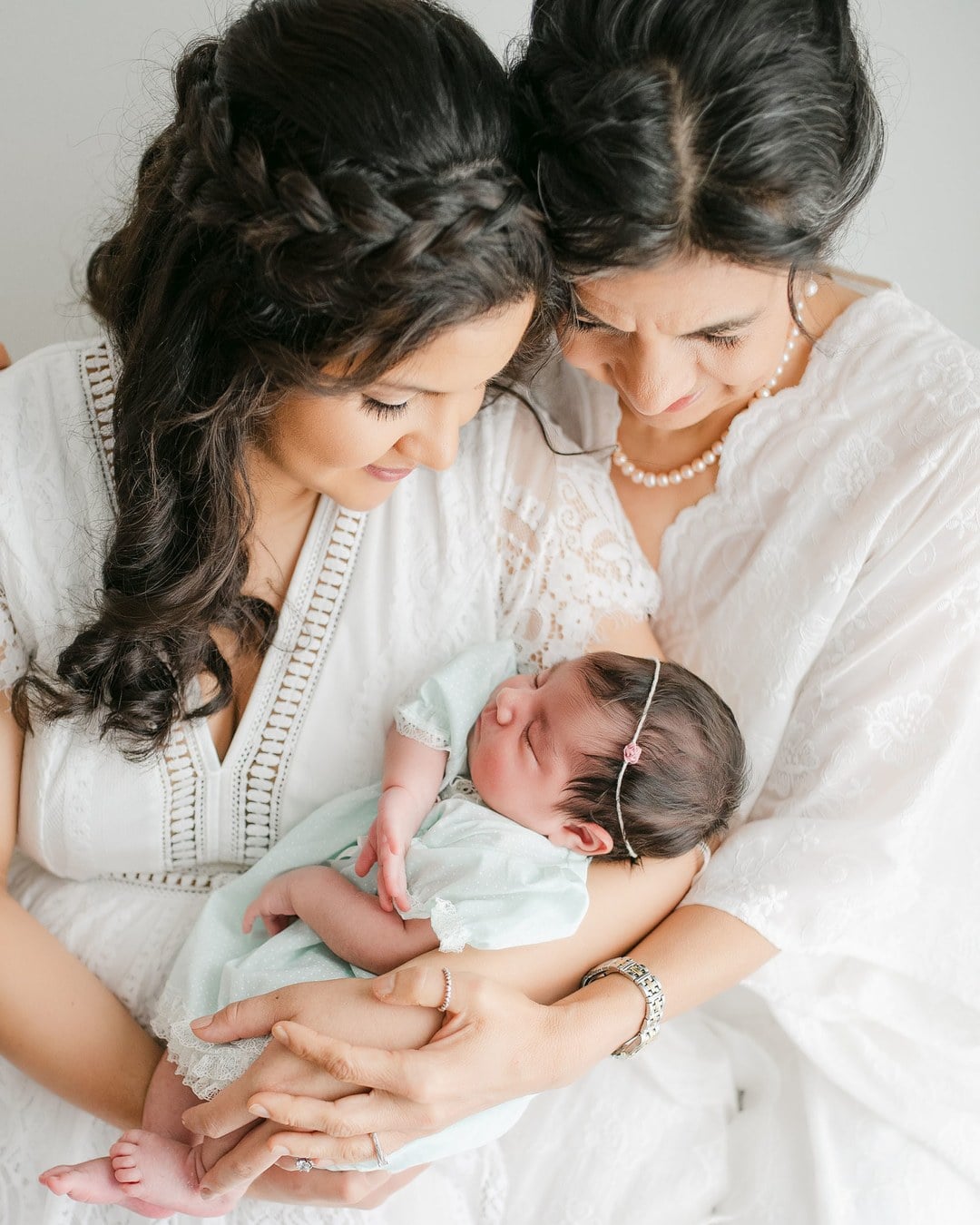 A mother and an older woman in white lace dresses leaning their heads together over a sleeping newborn in a mint outfit and floral headband