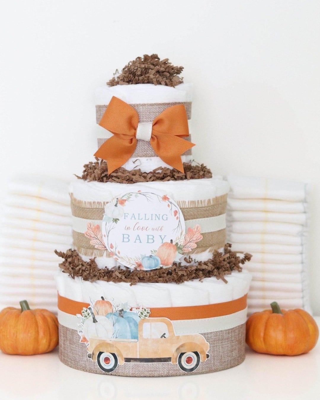 Three-tier diaper cake wrapped in burlap and orange ribbon with a tag and two small pumpkins beside it