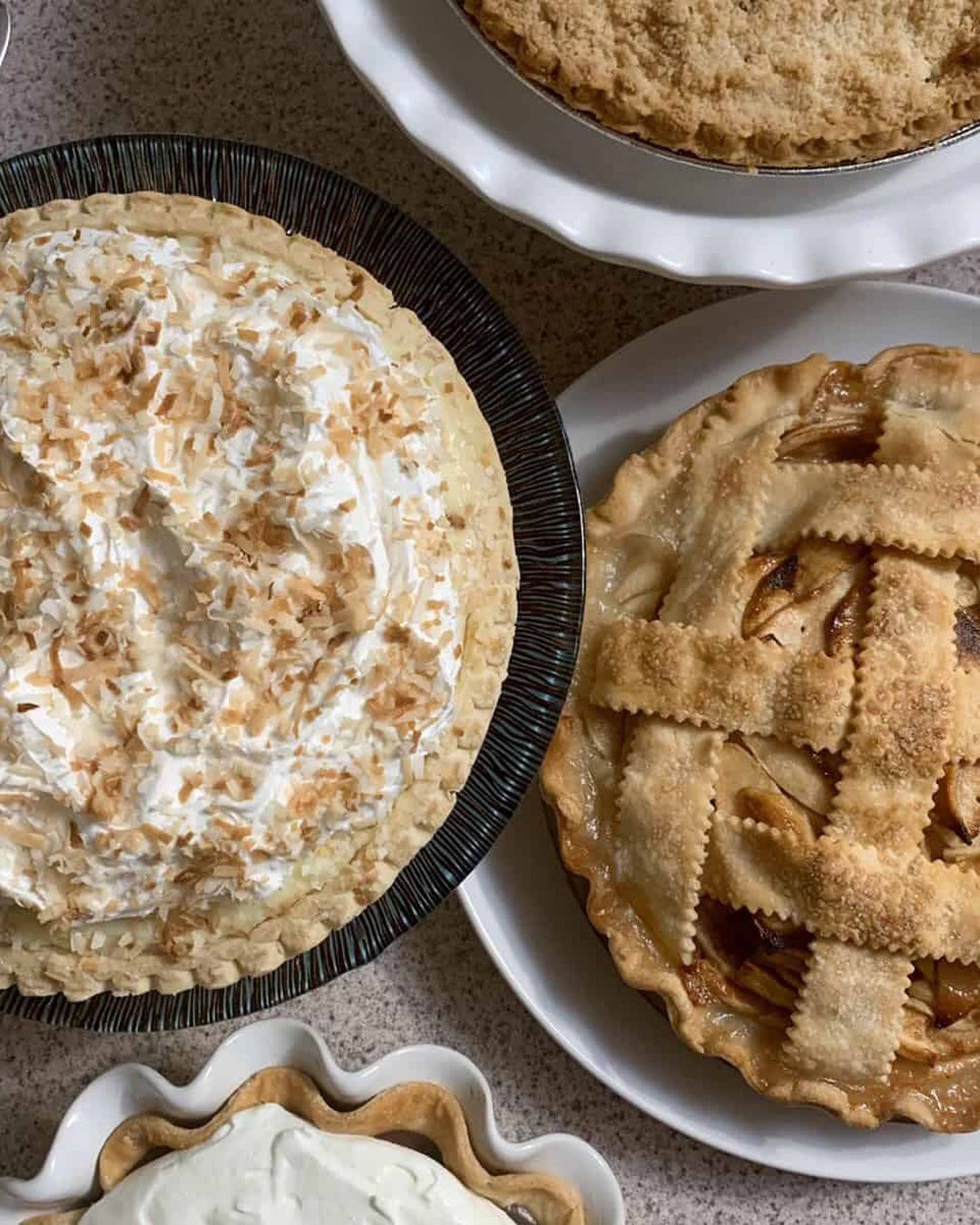 Overhead view of several homemade pies including coconut cream, lattice apple and crumb-topped on a speckled countertop