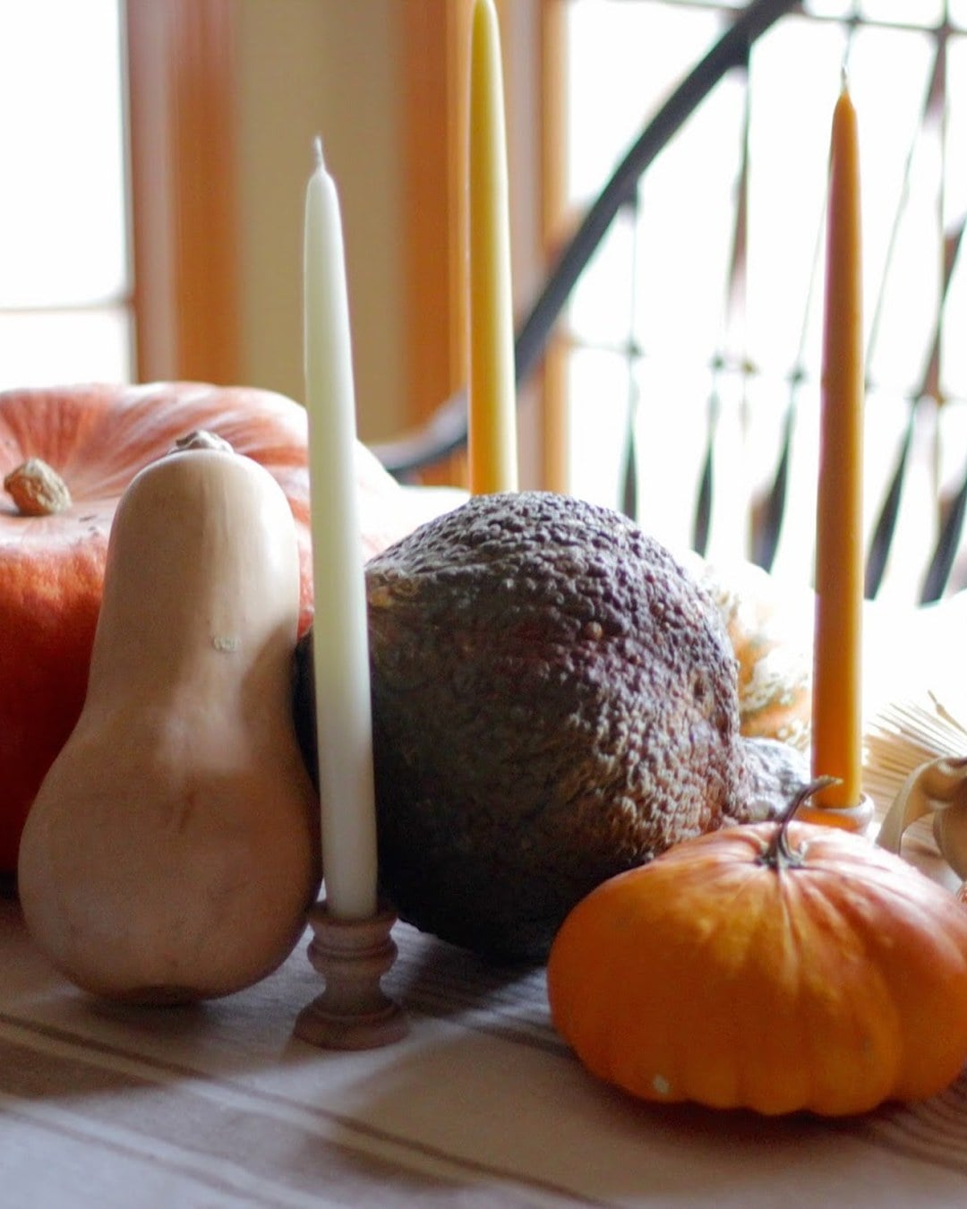 Table centerpiece with an orange pumpkin, butternut squash, warty gourd, wheat and tall taper candles on a striped linen runner