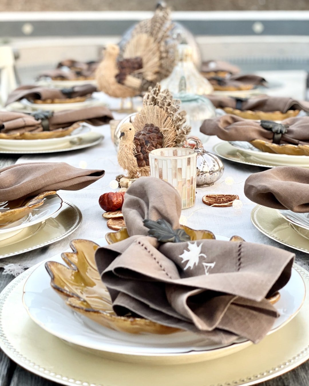Outdoor Friendsgiving table with mums in rusted milk cans, pumpkins, copper votives and cozy blankets on the benches