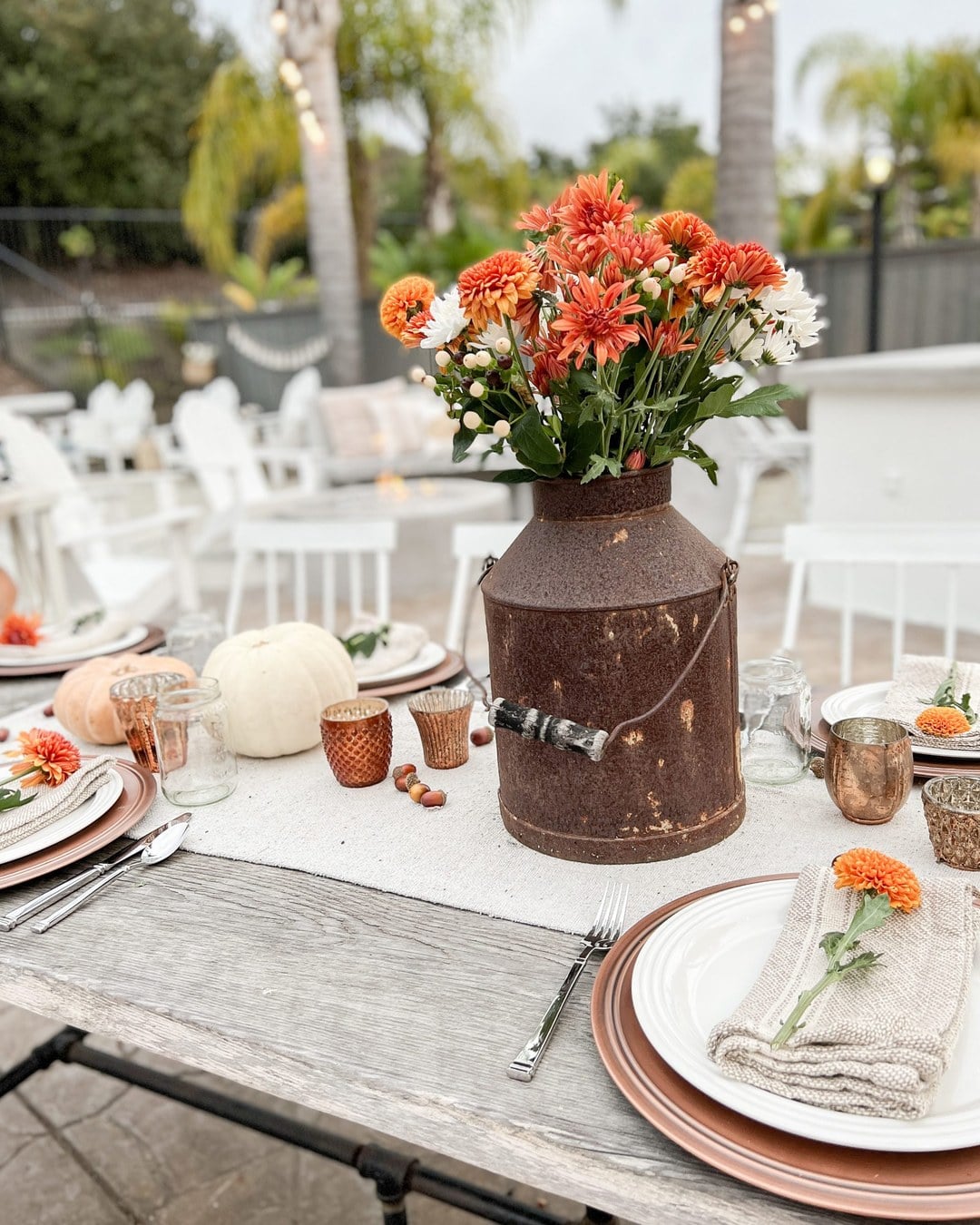 Rustic outdoor table with a white runner, orange and white mums in a milk-can vase, pumpkins, copper chargers and white plates
