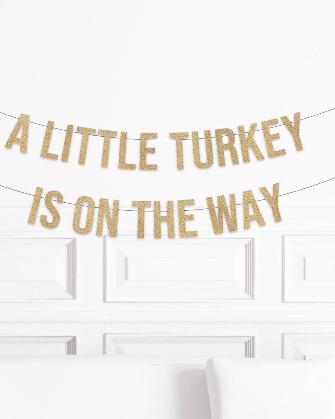 Gold glitter banner reading A LITTLE TURKEY IS ON THE WAY strung across a white paneled wall