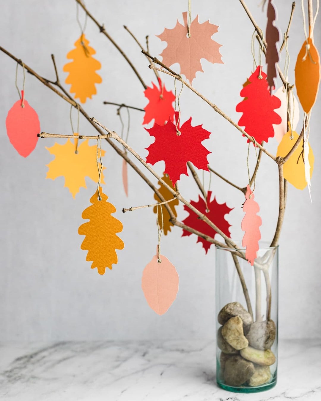Bare branches in a glass vase filled with stones, hung with colorful autumn paper leaves on twine