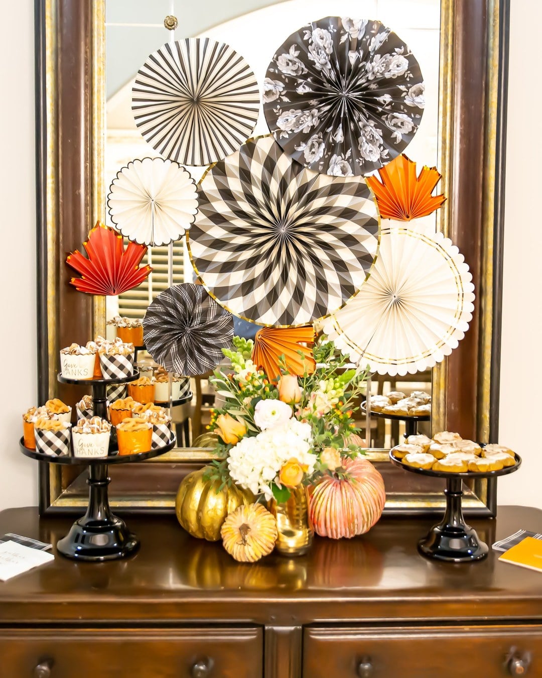 Wooden sideboard decorated with paper fans, a pumpkin centerpiece and cake stands holding Give Thanks cupcakes and cookies